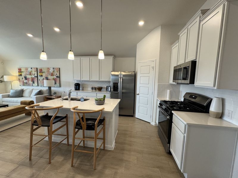 A modern kitchen with sleek white cabinets, a central island, and warm wood accents, opening into a cozy living area.