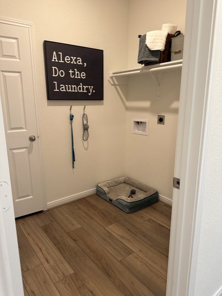 A cozy laundry room with wood flooring, wall decor, dog bed, and organized shelving. Perfect for functional living.