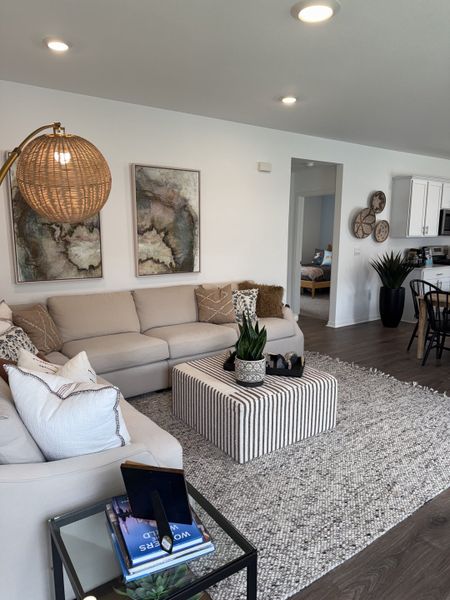 A cozy living room with a plush sectional, abstract art, a striped ottoman, and a contemporary woven lamp.