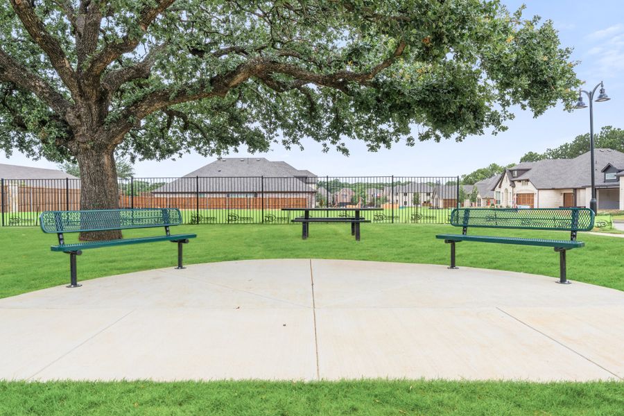 Neighborhood park seating area with green metal benches, picnic table, large shade tree, and iron fence bordering residential homes. Neighborhood park seating area with green metal benches, picnic table, large shade tree, and iron fence bordering residential homes.