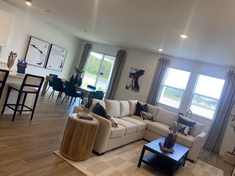 A cozy living space with a sectional sofa, wood floors, abstract art, and a bright dining area adorned with large windows.