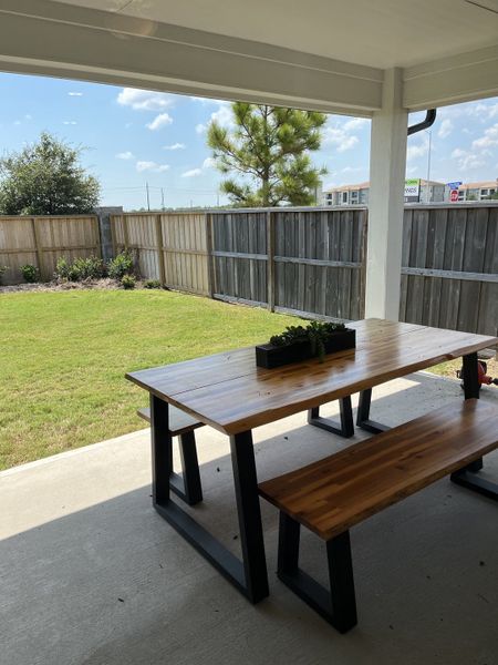 A cozy patio with a wooden picnic table, overlooking a neatly fenced backyard and lush green lawn.