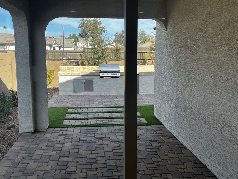 Covered patio leading to a built-in outdoor grill and well-maintained backyard with green artificial turf and pavers.