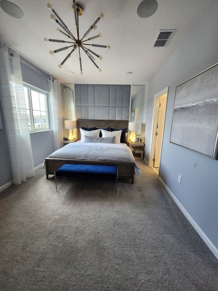 A modern bedroom with a statement chandelier, elegant panel headboard, plush carpet, and stylish decor in soft grays and blues.