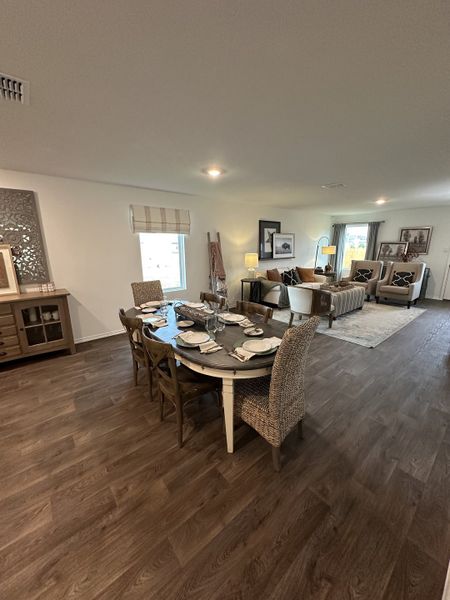 A cozy dining area with a rustic wooden table, woven chairs, and elegant decor leading into a bright, inviting living room.