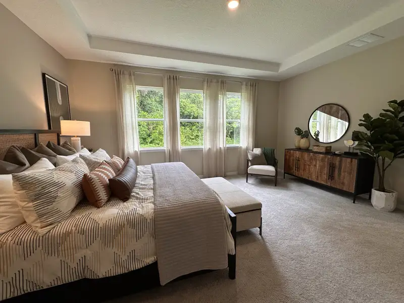A serene bedroom with a cozy bed, stylish armchair, and a wooden dresser, featuring soft lighting and lush green views.