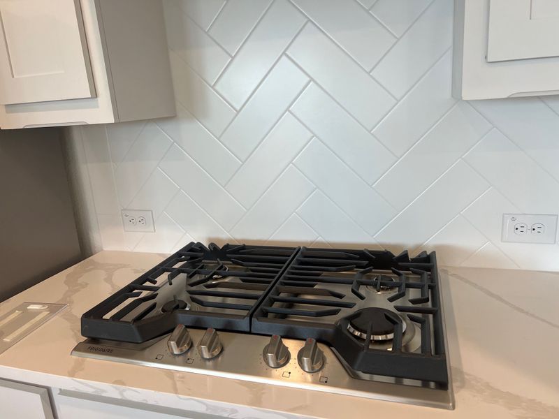 A modern kitchen with a sleek gas cooktop, white herringbone backsplash, and elegant marble countertops. A modern kitchen with a sleek gas cooktop, white herringbone backsplash, and elegant marble countertops.