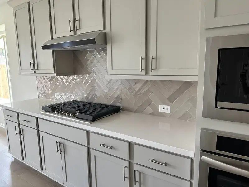 A modern kitchen featuring a sleek cooktop, herringbone tile backsplash, and elegant grey cabinetry. A modern kitchen featuring a sleek cooktop, herringbone tile backsplash, and elegant grey cabinetry.