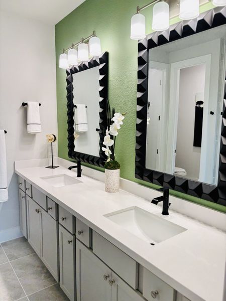 A chic bathroom featuring dual sinks, a green accent wall, geometric mirrors, and elegant lighting fixtures.