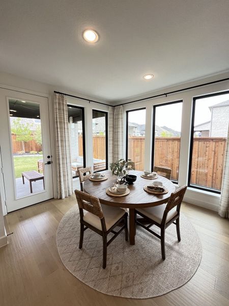 A cozy dining area with a round wooden table, woven chairs, and floor-to-ceiling windows overlooking a fenced backyard.