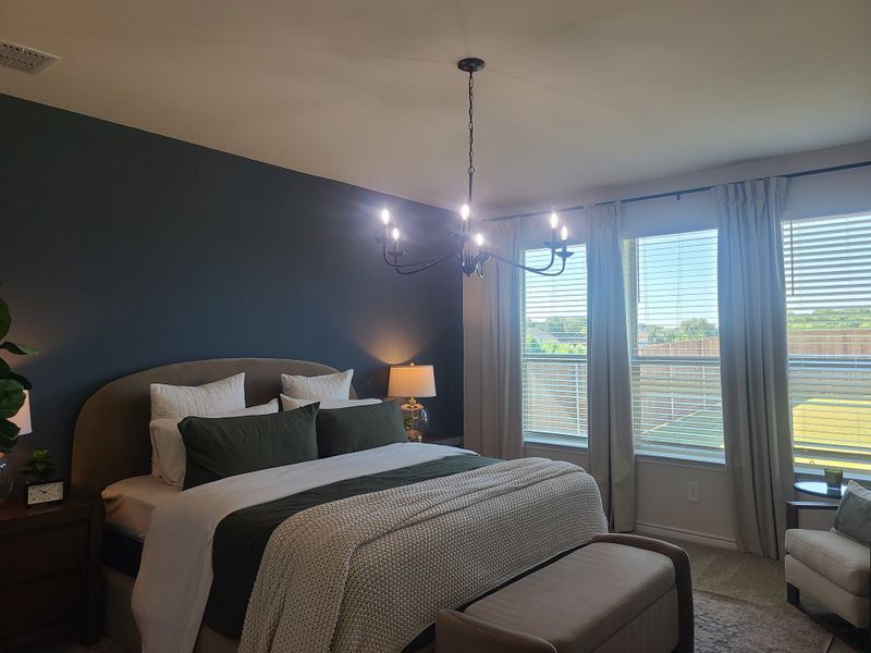 A cozy bedroom with a plush bed, dark accent wall, elegant chandelier, and large windows offering natural light.