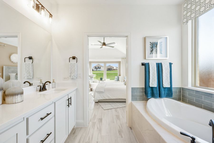 Ensuite bathroom with soaking tub, white double vanity, and open view into master bedroom with ceiling fan