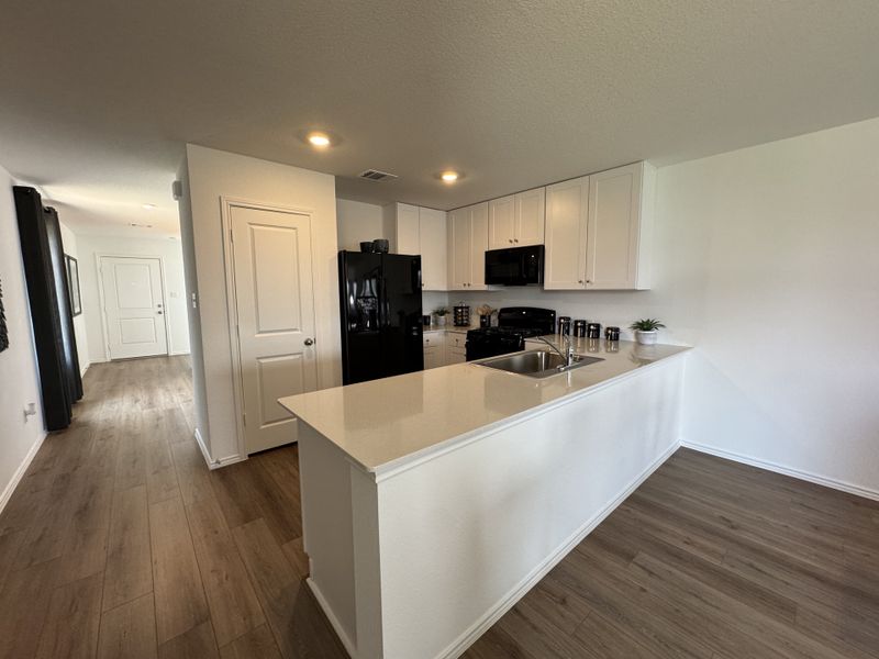 A modern kitchen with sleek white cabinets, black appliances, and wood flooring, leading to a bright, airy hallway.