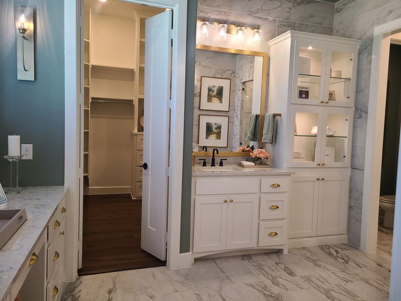 Elegant bathroom with marble tile, white cabinetry, gold accents, and soft lighting, leading to a spacious walk-in closet. Elegant bathroom with marble tile, white cabinetry, gold accents, and soft lighting, leading to a spacious walk-in closet.