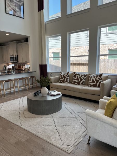 A modern living room with high ceilings, large windows, neutral-toned furniture, and an open kitchen layout.