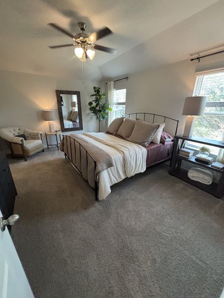A cozy bedroom with a ceiling fan, soft carpet, elegant bed, large mirror, and comfortable seating area by the window.