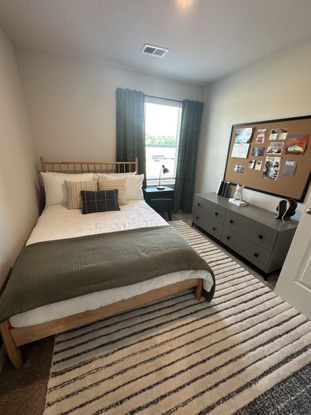 A cozy bedroom with a simple bed, striped rug, and a large corkboard for personal touches.