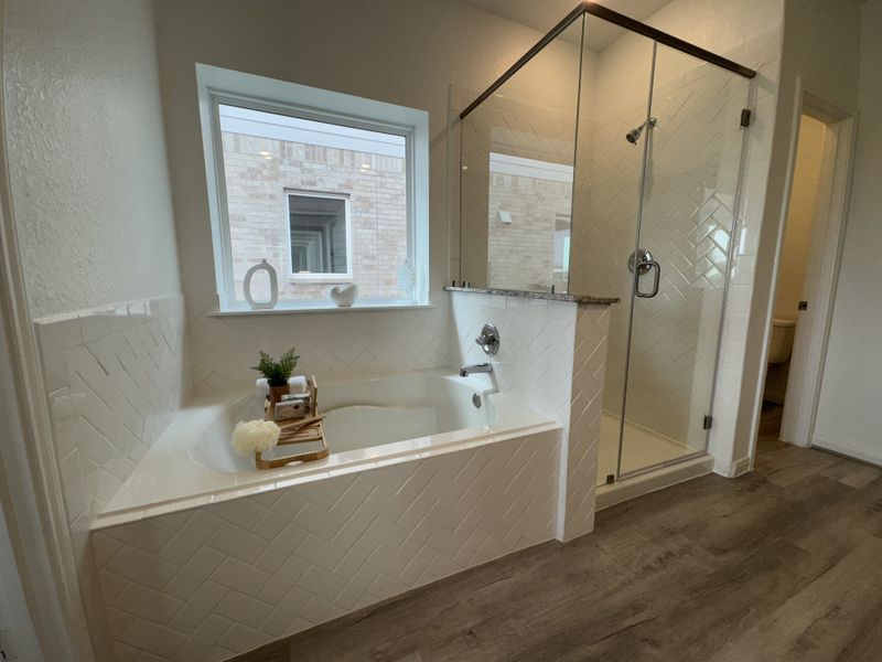 A modern bathroom featuring a glass shower, herringbone-tiled bathtub, and natural wood flooring.