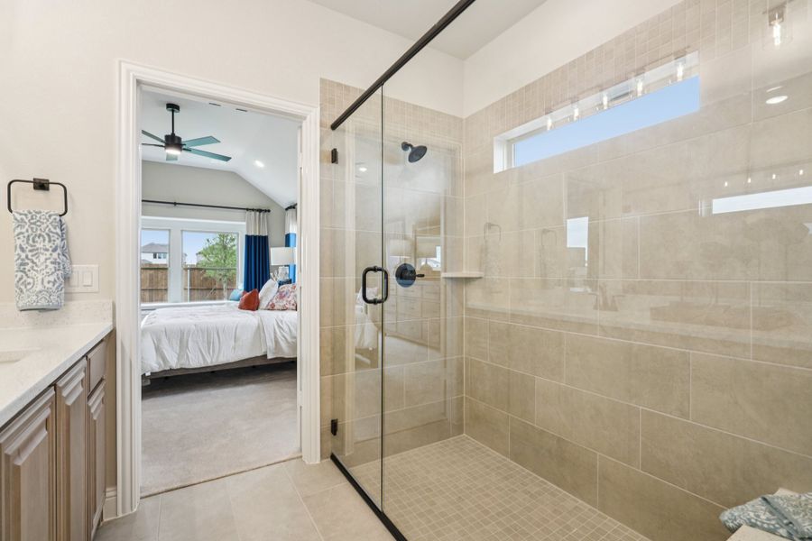 Ensuite bathroom with glass walk-in shower, beige tile walls, and view into master bedroom with ceiling fan Ensuite bathroom with glass walk-in shower, beige tile walls, and view into master bedroom with ceiling fan
