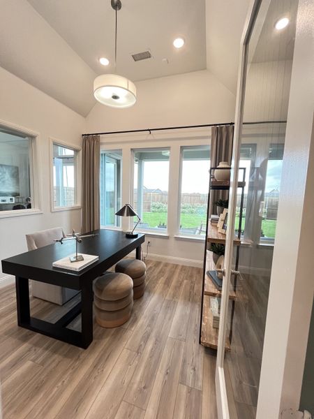 A modern office with elegant wood flooring, large windows, a sleek black desk, and stylish lighting.