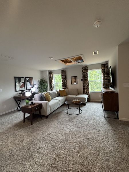 A cozy living room featuring a sectional sofa, carpeted floor, and natural light from large windows with patterned curtains.