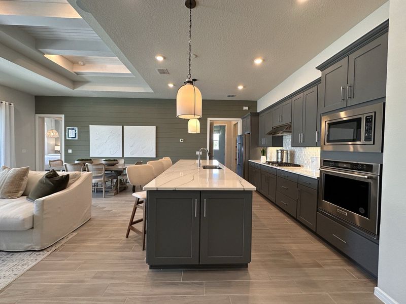 A modern kitchen with dark cabinets, an island, elegant lighting, and an open layout leading to a dining area.