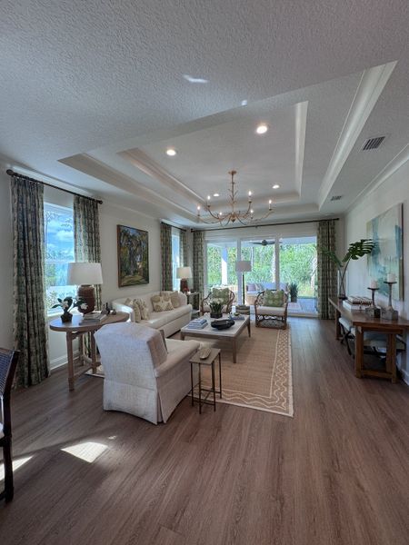 A bright, elegant living room with a tray ceiling, refined chandelier, and floor-to-ceiling windows overlooking greenery.