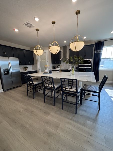 A grand kitchen with dark cabinetry, gold pendant lighting, and a spacious island, seamlessly flowing into the open living area in Crossroads at Kelly Park by Dream Finders Homes (Apopka, FL).

