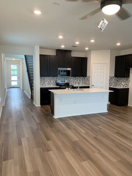 Modern kitchen with dark cabinets, mosaic backsplash, island, and wood flooring leading to a bright hallway.