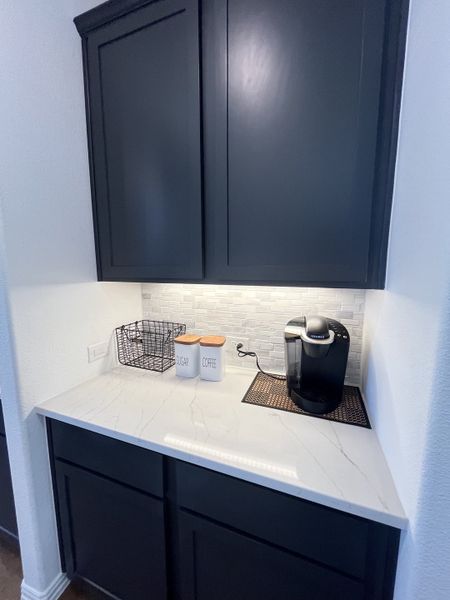 A cozy coffee nook with dark cabinetry, elegant marble countertop, and chic white backsplash.