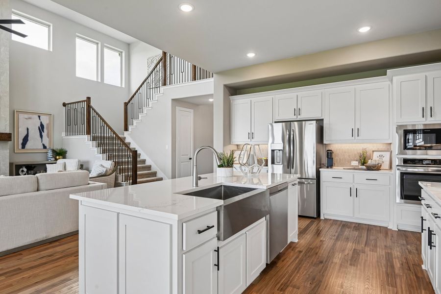 Open-concept kitchen with white cabinets, large island, stainless steel appliances, and hardwood floors overlooking living area and staircase. Open-concept kitchen with white cabinets, large island, stainless steel appliances, and hardwood floors overlooking living area and staircase.
