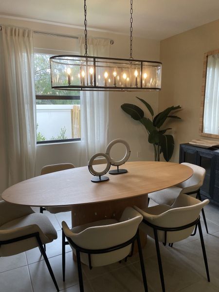 A serene dining area with an oval wooden table, modern chairs, and an elegant chandelier, alongside a large decorative plant.