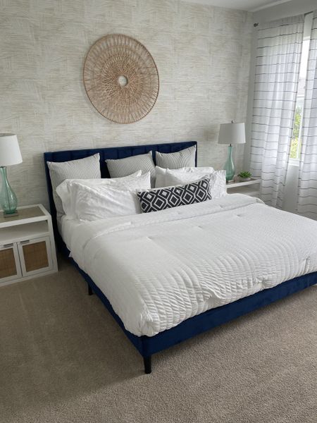 A cozy bedroom featuring a plush navy bed, textured wall decor, modern lamps, and sheer curtains for a bright ambiance.