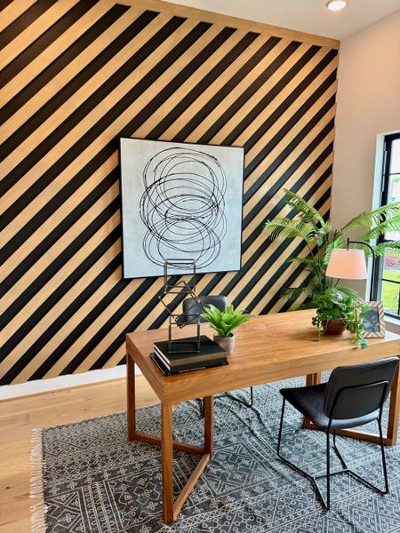 A chic home office with a diagonal wood accent wall, modern art, wood desk, and lush plants on a patterned rug.