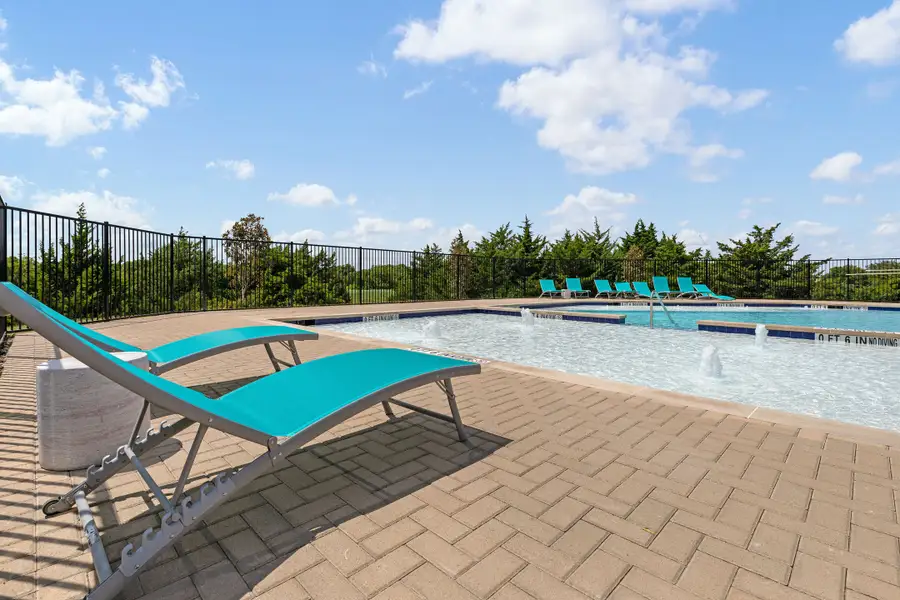 Outdoor community pool with water features, teal lounge chairs, brick patio, and black iron fence