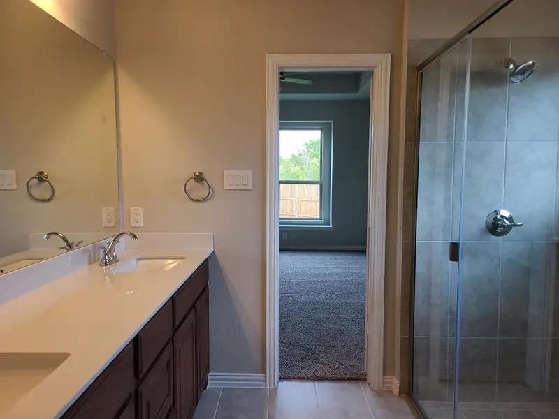 Modern bathroom featuring a sleek shower, dual vanity, and large mirror, leading to a carpeted room with a large window.