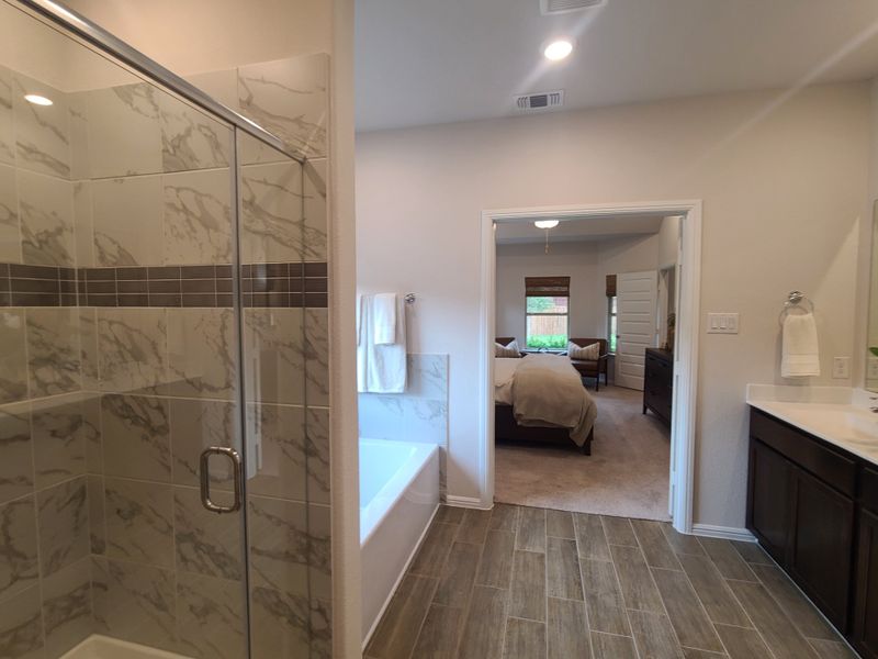 A luxurious bathroom with a marble-tiled shower, elegant tub, wood-patterned flooring, and double vanity leading to a cozy bedroom. A luxurious bathroom with a marble-tiled shower, elegant tub, wood-patterned flooring, and double vanity leading to a cozy bedroom.