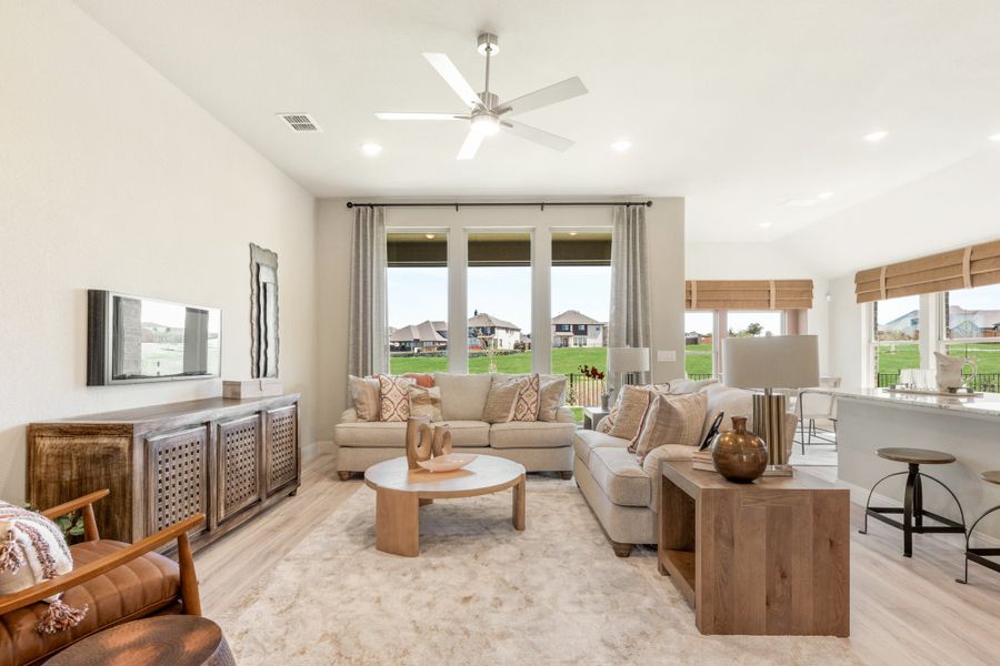 Bright living room with beige sofas, ceiling fan, and large windows overlooking a green yard