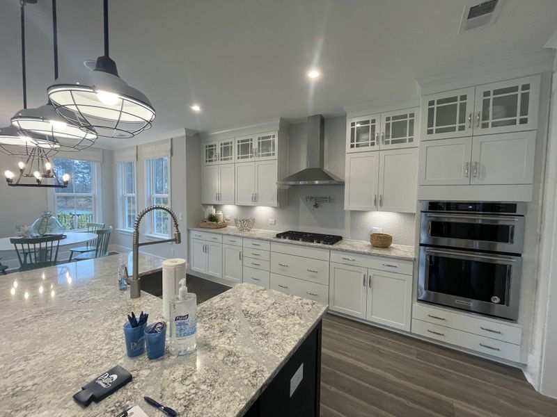 A bright, modern kitchen with white shaker cabinets, double ovens, a gas cooktop, and a spacious granite island with industrial pendant lights.