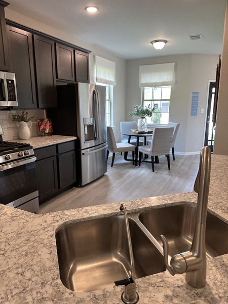 A modern kitchen with dark cabinets, stainless appliances, and a cozy dining area, featuring bright windows and light floors.