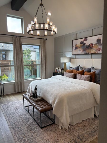 A cozy bedroom featuring a plush bed, elegant chandelier, large windows, and tasteful art, with a welcoming ambiance.