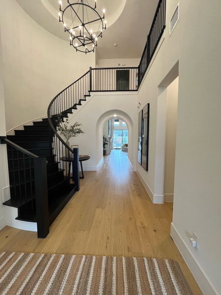 A grand foyer with a sweeping black staircase, wooden floors, and elegant chandelier lighting.