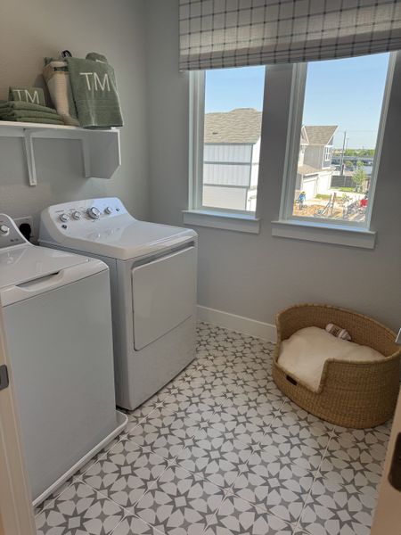 A bright laundry room with patterned tiles, modern washer and dryer, and a cozy pet bed by the window.