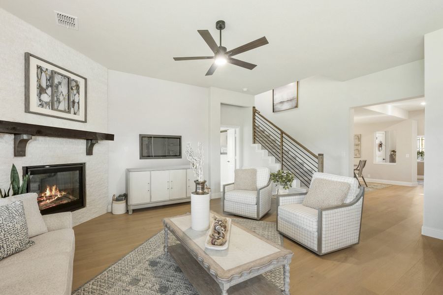 Living room with white brick fireplace, hardwood floors, ceiling fan, and staircase in background