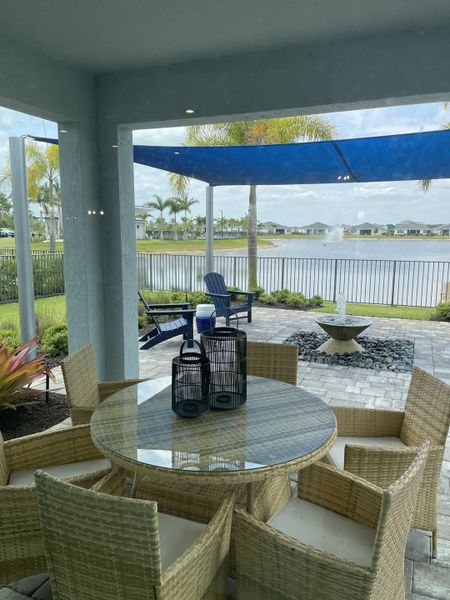 A serene patio with wicker seating, glass table, and lake view, featuring a fire pit and relaxing blue shade sail. A serene patio with wicker seating, glass table, and lake view, featuring a fire pit and relaxing blue shade sail.