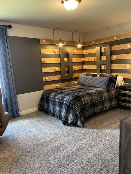 A cozy bedroom with a plaid bedspread, wood accent wall featuring large initials, and warm pendant lighting.