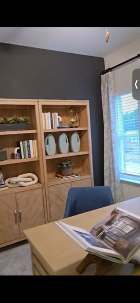 A cozy home office with wooden bookshelves, dark accent wall, and natural light through a window with elegant curtains.