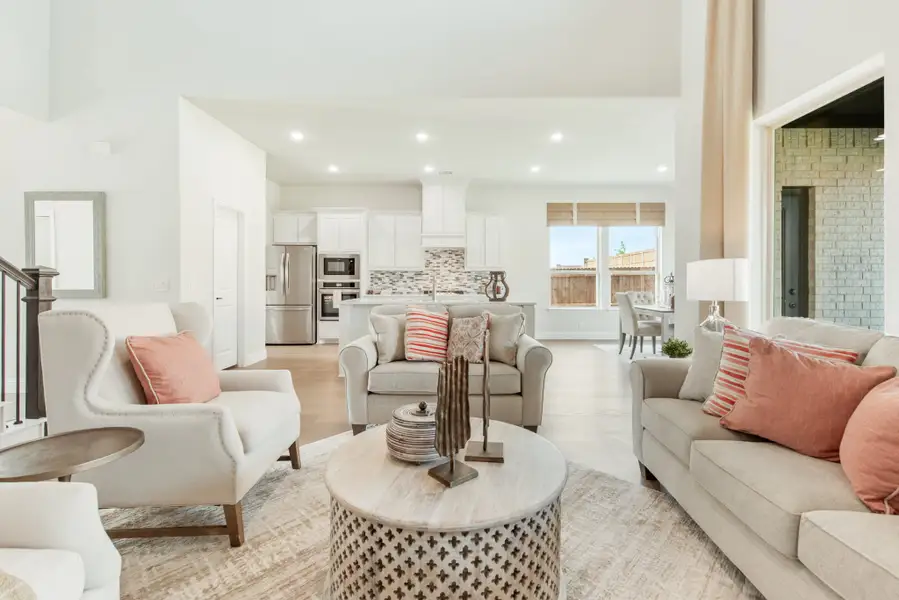 Open-concept living room with neutral seating and pink accent pillows, flowing into a white kitchen with stainless steel appliances