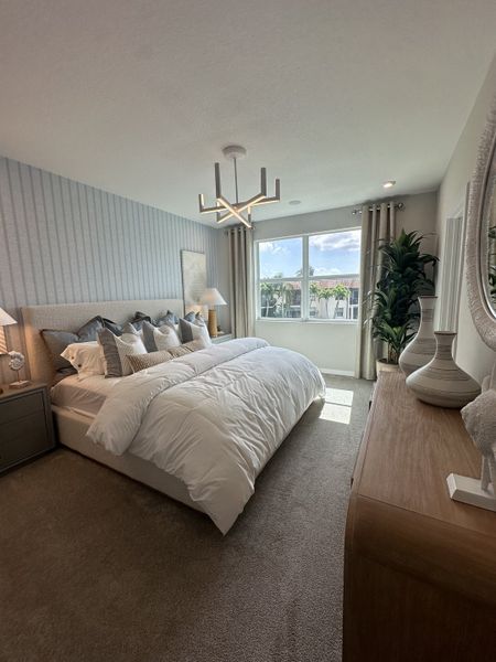 A cozy bedroom with a plush bed, modern chandelier, and large window offering a view, styled with neutral tones and greenery.