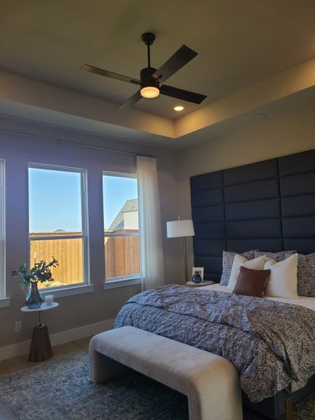 A cozy bedroom with a plush bed, modern ceiling fan, and large windows bringing in natural light.